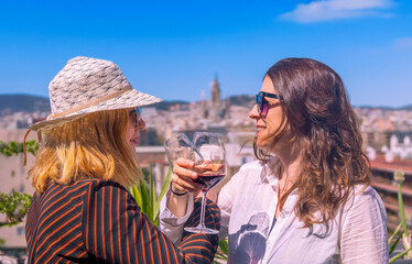 A happy lesbian couple toasting with red wine, their glasses crossed, looking at each other on a sunny rooftop overlooking the city skyline and Barcelona's Sagrada Familia.