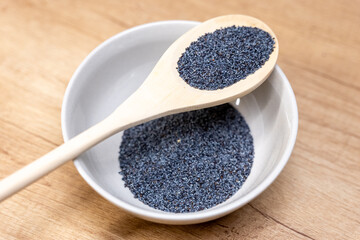 A wooden spoon filled with blue-gray poppy seeds held above a bowl of poppy seeds on a wooden surface, shown up close for cooking, baking, nutrition, and ingredient use.