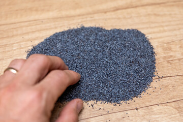 A hand touching a pile of blue-gray poppy seeds on a wooden surface, shown in a close-up scene for cooking, baking, nutrition, and ingredient handling concepts.