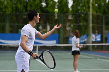 A Male tennis player tosses the ball to serve while another player waits in the background, capturing a natural training moment on the court