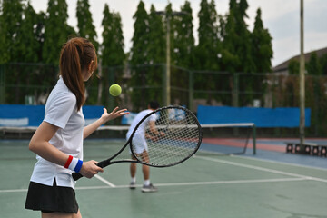 An outdoor tennis practice session captures a young female player tossing a serve while her teammates wait to receive it. Team tennis