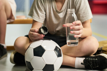 Caucasian child sitting on floor holding plastic water bottle and cap with soccer ball resting against legs, showing hands and lower body, preparing for sports activity indoors