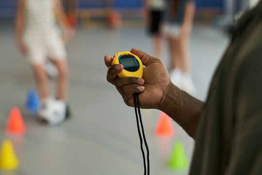 Black man holding stopwatch timing group of children participating in physical activity indoors, blurred kids and colorful cones visible in background, focus on hand and device