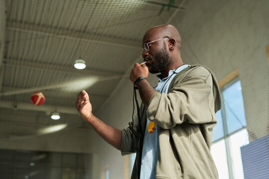 Black man standing in gymnasium blowing whistle and gesturing with hand, appearing to coach or instruct during sports activity, basketball hoop visible in background