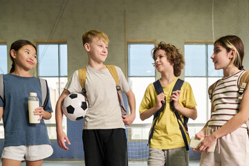 Group of four children including Asian girl, Caucasian boy holding soccer ball, Caucasian boy and Caucasian girl standing together smiling and talking in indoor sports facility