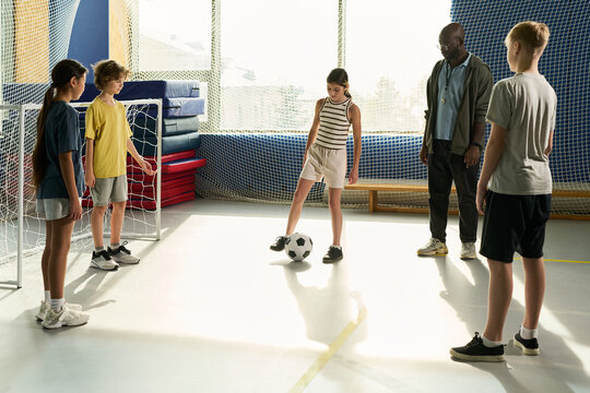Group of multiethnic teenagers standing in gymnasium playing soccer, female teenager controlling ball with foot while male coach observing, sunlight streaming through window