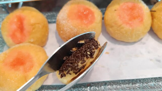 Close-up of soft sweet bread topped with chocolate sprinkles, displayed on a serving tray. The bread has a golden-yellow color with sugar coating and fruit jam center, commonly served as bakery snacks