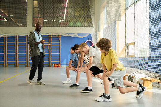 Black man coaching group of children stretching legs during physical education class in gymnasium, children performing lunges while listening to instructions, sports equipment visible in background