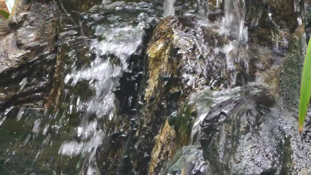 Close-up of clear water flowing over moss-covered rocks in a small garden waterfall. Natural stream movement creating relaxing and calming atmosphere. Suitable for nature background, tranquility conce