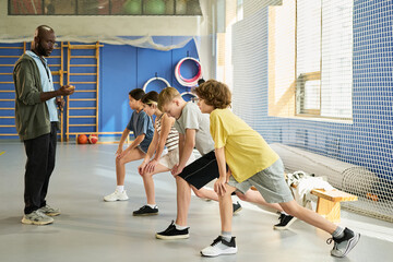 Black man instructing group of four teenagers stretching in gym, teenagers preparing for physical activity, sports equipment visible in background, coach leading warm up