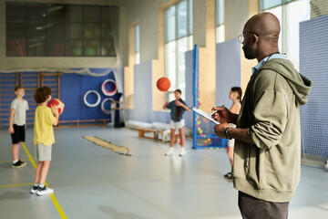 Black man supervising group of teenagers practicing basketball in gymnasium, holding clipboard and observing students dribbling and passing basketballs during physical education class