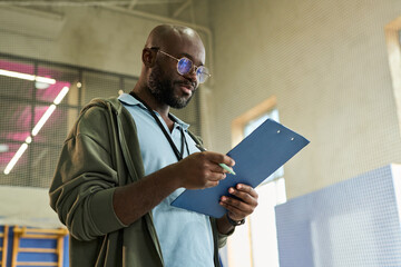 Young Black man wearing glasses standing indoors holding clipboard and writing with pen, appearing focused while reviewing notes or making checklist in professional environment