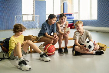 Group of four children sitting on gym bench, two girls and two boys, multiethnic, interacting with smartphone and holding basketball and soccer ball, relaxed after sports activity