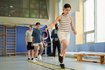 Obraz premium Group of multiethnic children practicing agility drills in gym, leading Caucasian girl balancing on agility ladder while classmates and Black man coach observing and supervising activity