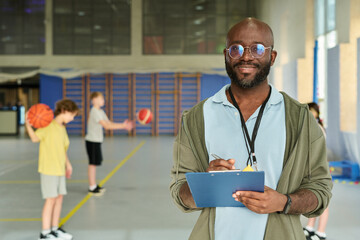 Portrait of Black man smiling and holding clipboard while standing in gymnasium, teenagers playing basketball in background, coach supervising youth sports activity