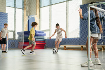 Group of Caucasian and multiethnic children playing soccer indoors, girl dribbling ball while boys and girl defending goal, energetic movement in gymnasium with sports equipment visible