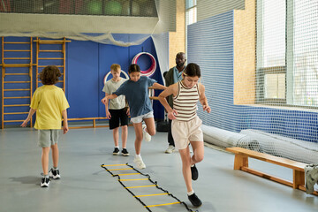 Group of multiethnic children exercising in gym, jumping over agility ladder while Black man supervising activity, kids demonstrating coordination and teamwork during physical education class