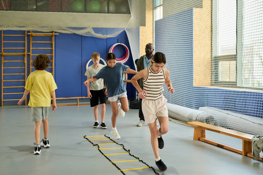 Group of multiethnic children exercising in gym, jumping over agility ladder while Black man supervising activity, kids demonstrating coordination and teamwork during physical education class