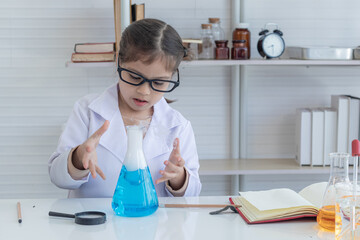 Cheerful Arabian student girl wearing eyeglasses using magnifier glass looking smoke liquid experiment in flask, pupil kid in lab coat uniform having fun attention to cylinder test, a future scientist