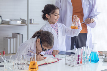 Adorable asian girls wearing lab coat whispering secrets and making experiment in chemistry class, little children whisper excitedly while science experiment together in classroom.