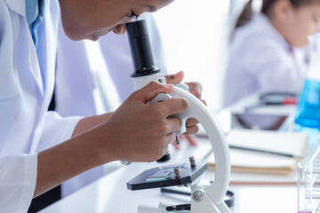 Close-up group of multicultural cute elementary students kids wearing lab coat uniforms using microoscope writing experiment report in laboratory room, lifestyle pupil boy and girls learning education