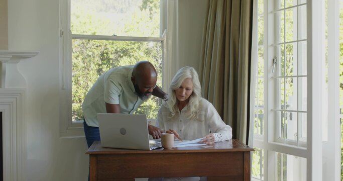 Reviewing couple leaning over wooden desk at home, pointing at paperwork next to laptop - Powered by Adobe