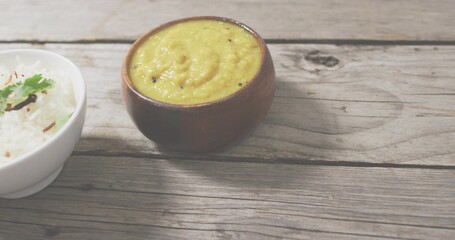 Serving brown clay bowl holding yellow dal curry on rustic wood tabletop, with white ceramic bowl