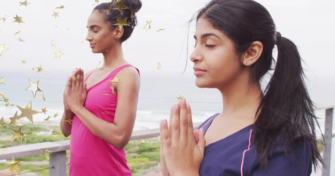Meditating woman in navy athletic top placing palms on deck facing dunes and ocean, gold stars - Powered by Adobe