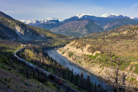 Long cargo train moving along Thompson River with snow capped mountain
