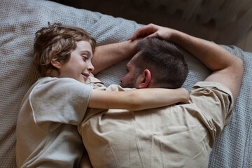 Lifestyle photo of boy and his bearded dad lying on bed and resting. Son hugging father at home. Family value, heart-to-heart conversations, father's day.