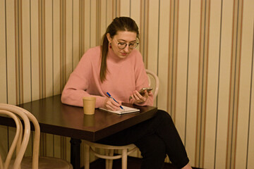 A young woman in a pink sweater sits at a cafe table and writes attentively in her notebook. The angle is close, showing her focused expression and hand movements.