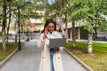 Young african american businesswoman walking and managing remote work, talking on phone while using laptop in a park