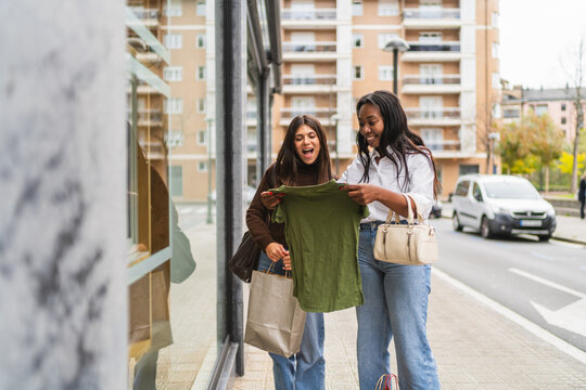 Two diverse women friends shopping, excitedly looking at a green t shirt near a store window on a city street - Powered by Adobe