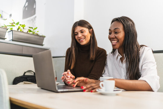 Two smiling multi ethnic women friends enjoying coffee and working together on a laptop in a modern cafe