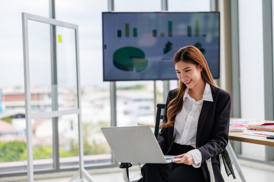 Focused Asian professional woman sitting at a desk, confidently working on digital tasks using both a modern laptop and a touch screen tablet. Capturing concepts of remote work, technology, innovation - Powered by Adobe