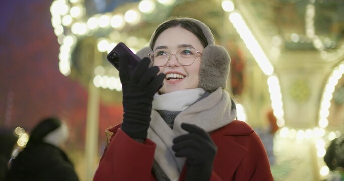 A young beautiful girl in a red coat walks in the evening at the Christmas fair and talks on the phone or records a voice message standing against the backdrop of a carousel with illuminations