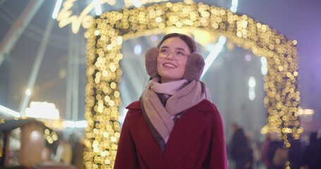 A young beautiful girl in a red coat walks in the evening at the New Year's Christmas fair against the backdrop of a Ferris wheel among lights and garlands and admires the decorations
