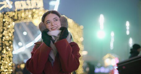 A young beautiful girl in a red coat walks in the evening at the New Year's Christmas fair against the backdrop of a Ferris wheel among lights and garlands and admires the decorations