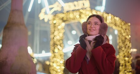 A young beautiful girl in a red coat walks in the evening at the New Year's Christmas fair against the backdrop of a Ferris wheel among lights and garlands and admires the decorations