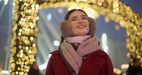 A young beautiful girl in a red coat walks in the evening at the New Year's Christmas fair against the backdrop of a Ferris wheel among lights and garlands and admires the decorations