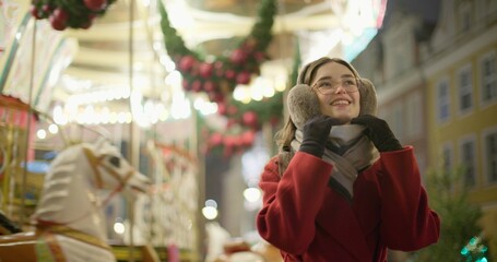 A young beautiful girl in a red coat walks in the evening at the European Christmas Market against the backdrop of a festive carousel with illuminations.