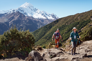 Smiling woman hiking with male companion on rocky mountain trail in Himalayas, lush green forest slope and snow mountain in background.