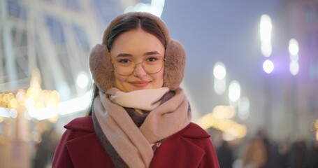 A young beautiful girl in a red coat walks in the evening at the New Year's Christmas fair against the backdrop of a Ferris wheel among lights and garlands and admires the decorations