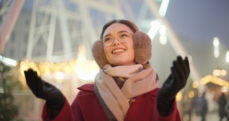A young beautiful girl in a red coat walks in the evening at the New Year's Christmas fair against the backdrop of a Ferris wheel among lights and garlands and admires the decorations