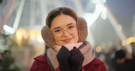 A young beautiful girl in a red coat walks in the evening at the New Year's Christmas fair against the backdrop of a Ferris wheel among lights and garlands and admires the decorations