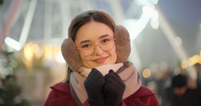 A young beautiful girl in a red coat walks in the evening at the New Year's Christmas fair against the backdrop of a Ferris wheel among lights and garlands and admires the decorations - Powered by Adobe