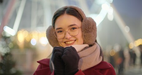 A young beautiful girl in a red coat walks in the evening at the New Year's Christmas fair against the backdrop of a Ferris wheel among lights and garlands and admires the decorations
