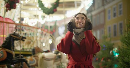 A young beautiful girl in a red coat walks in the evening at the European Christmas Market against the backdrop of a festive carousel with illuminations.