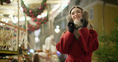 A young beautiful girl in a red coat walks in the evening at the European Christmas Market against the backdrop of a festive carousel with illuminations.