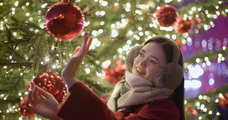 A young beautiful girl in a red coat stands near a Christmas tree with garlands and illuminations in the evening at the European Christmas Market and touches the Christmas tree decorations.